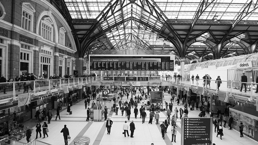 Black and white photo of a bustling train station interior with travelers and arrival/departure boards.