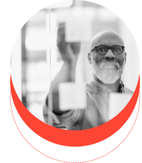 A smiling senior bald man with a beard, wearing glasses and holding a consumption metering device for engineers, framed within a red circular border, against a blurred office background.