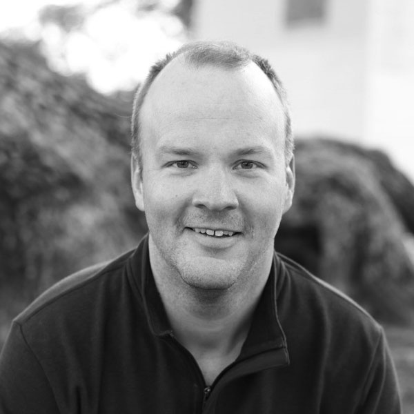 Black and white portrait of a smiling bald man wearing a polo shirt, sitting outdoors with blurred foliage in the background, taken at a Zuora Subscribed Live event.