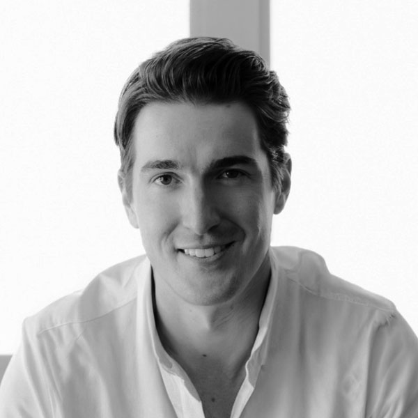 Black and white portrait of a smiling man with slicked-back hair, wearing a white shirt, sitting indoors with a bright background at the Zuora Subscribed Live event.
