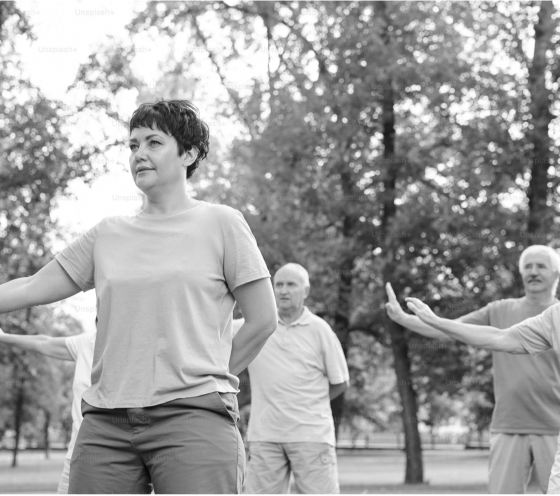 A black and white image of a middle-aged woman and two men practicing tai chi in a park with trees in the background.