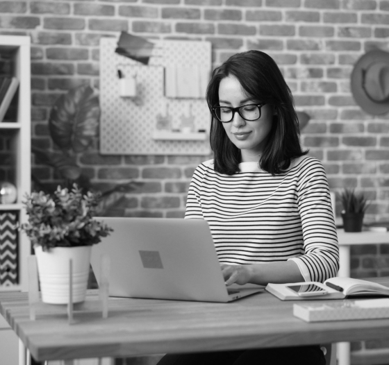 Person with glasses typing on a laptop at a desk with a brick wall background. Plant, books, and phone placed on the desk.