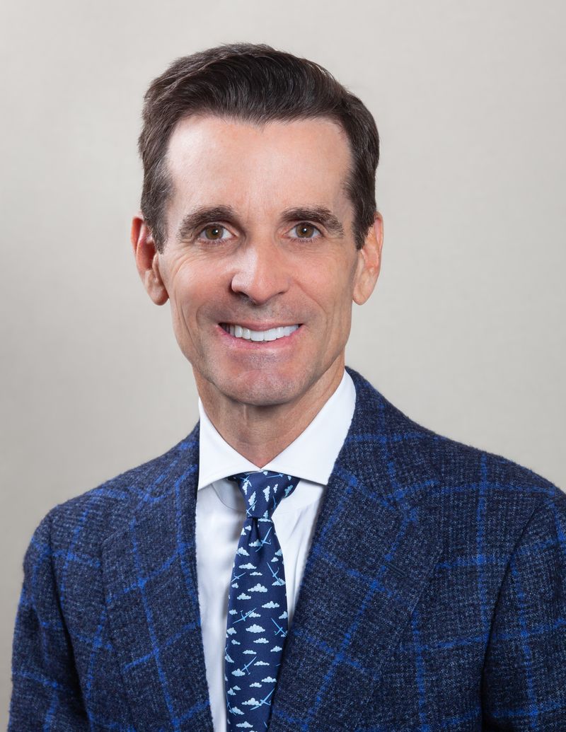 A professional portrait of a smiling man with dark hair, wearing a blue checked blazer and a tie with a bird pattern.