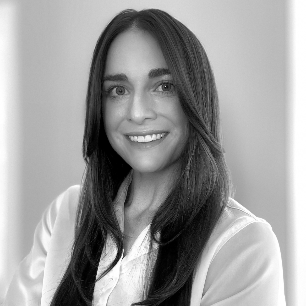 A woman with long dark hair wearing a white shirt is smiling at the camera in a black and white portrait.