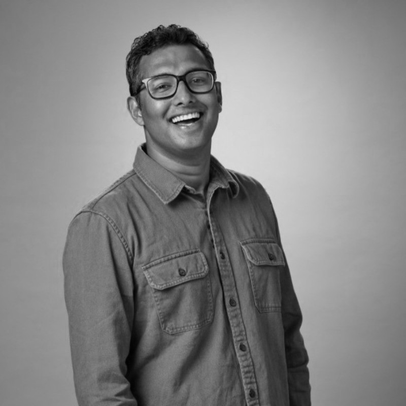 Black and white image of a person wearing glasses and a button-up shirt, smiling at the camera.