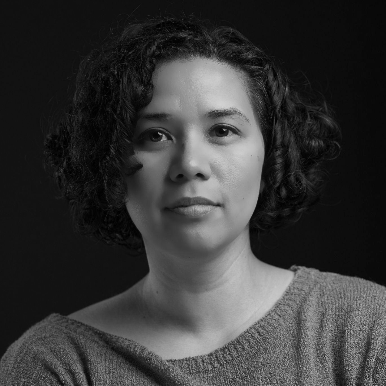 Black and white portrait of a person with short, curly hair, wearing a sweater, looking directly at the camera with a neutral expression.