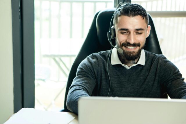 Man with a beard wearing a headset smiles while working on a laptop in a bright office setting.