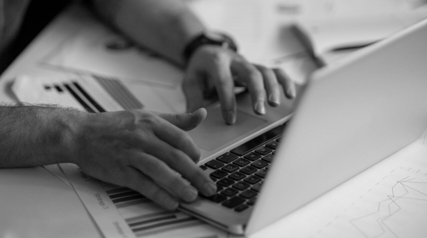 Two men looking at a laptop screen, with one typing, over a desk covered in papers. Black and white image.