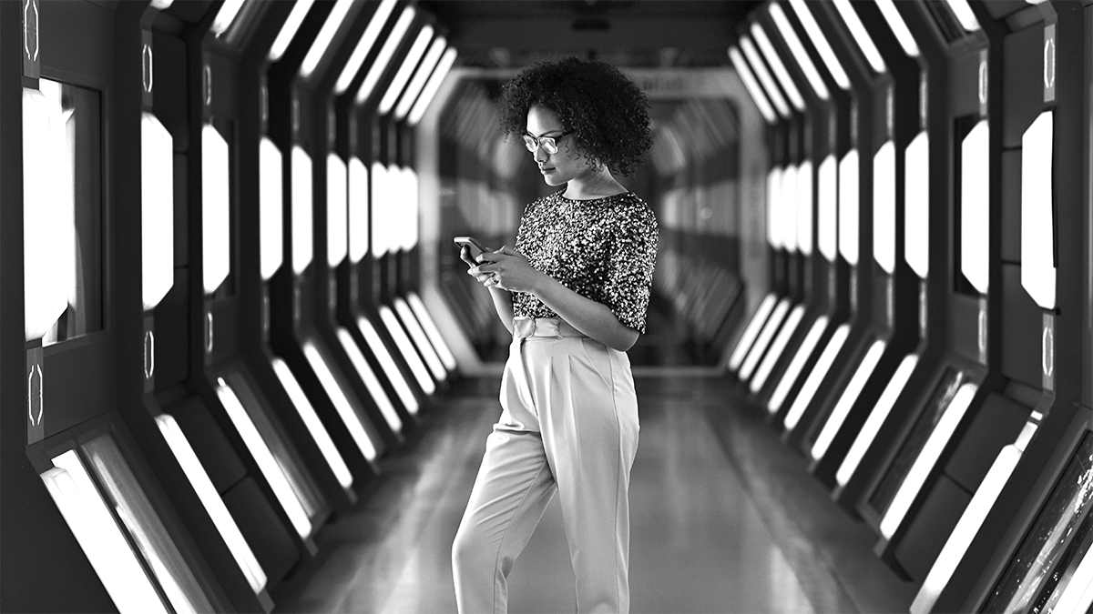 A person stands in a futuristic corridor, looking at their phone. The corridor is lined with illuminated panels.