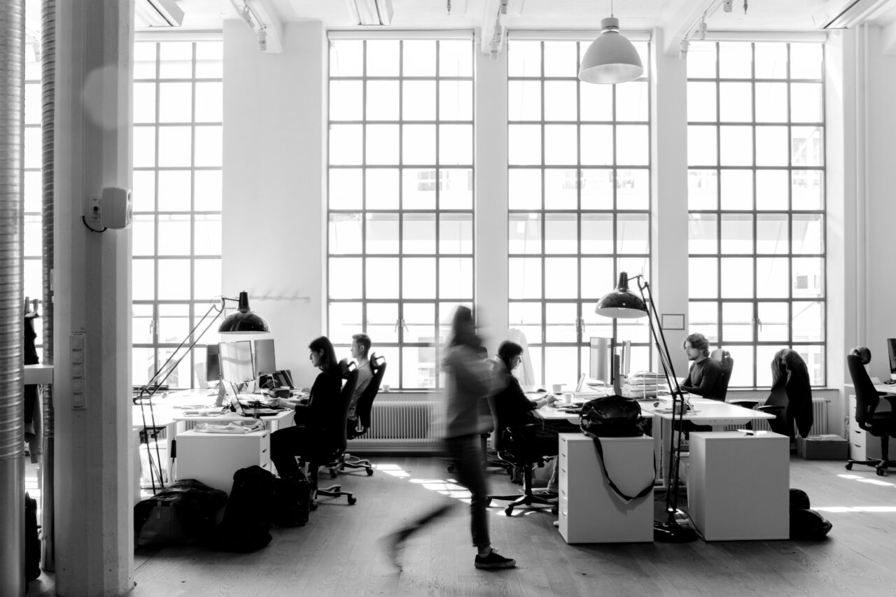 Black and white photo of an open office with several people working at desks, large grid windows in the background, and a person blurred while walking through the room.