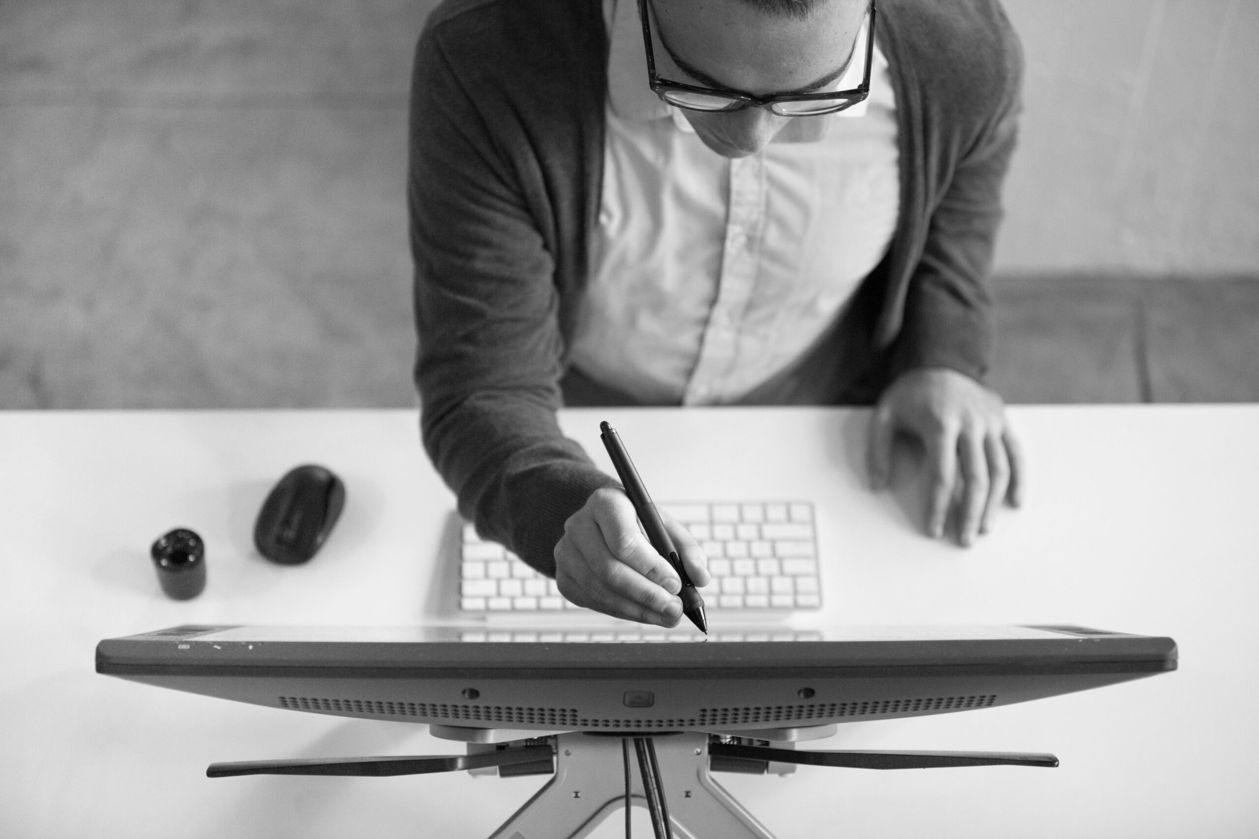 A person using a stylus to interact with a desktop monitor while working at a desk, viewed from above.