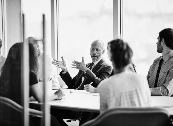 A man in a suit is speaking animatedly to a group of people seated around a conference table in an office setting.