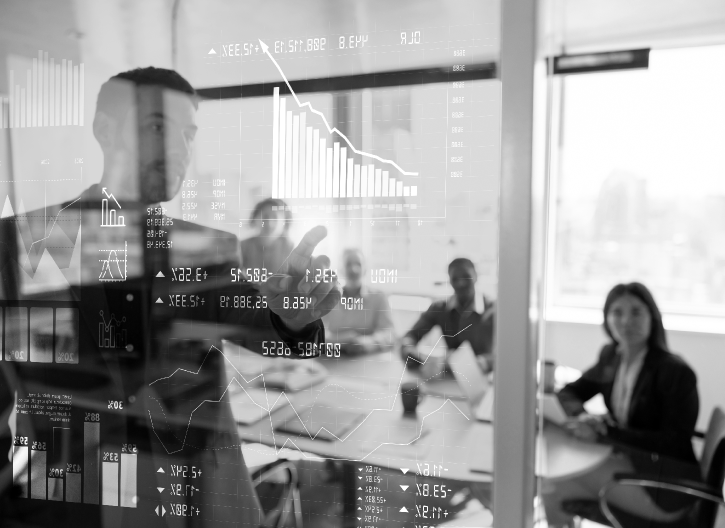 A man gestures towards a glass wall displaying various financial charts and graphs while three people seated at a table listen attentively in a bright office.