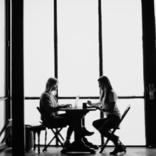 Two people sit at a table in front of large windows, using laptops. Their silhouettes are visible against the bright light.