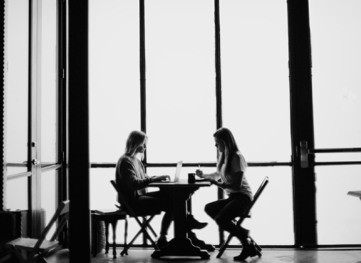 Two people sit at a table in front of large windows, using laptops. Their silhouettes are visible against the bright light.