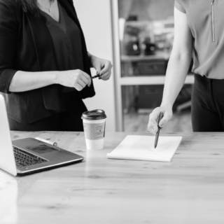 Two people stand around a table with a laptop, coffee cup, and document. One points at the paper with a pen while the other holds an object. The scene appears to take place in a professional setting.