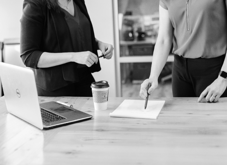 Two people stand around a table with a laptop, coffee cup, and document. One points at the paper with a pen while the other holds an object. The scene appears to take place in a professional setting.