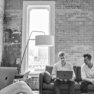 Three men sit on a couch and chairs with laptops and a coffee cup in a brick-walled room with large windows and a floor lamp.