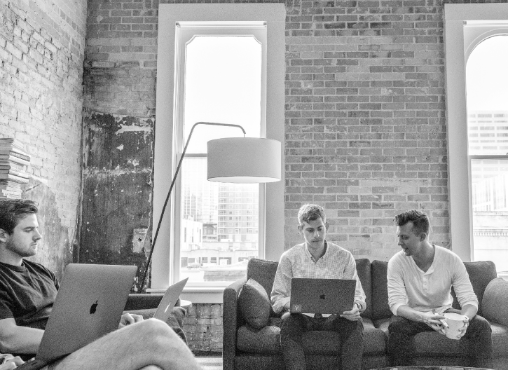 Three men sit on a couch and chairs with laptops and a coffee cup in a brick-walled room with large windows and a floor lamp.