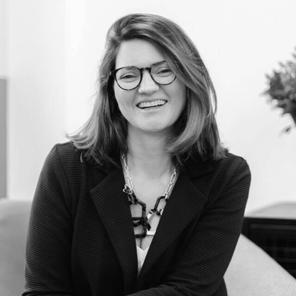 A woman with shoulder-length hair and glasses, wearing a dark jacket and a chunky necklace, smiling while seated indoors.