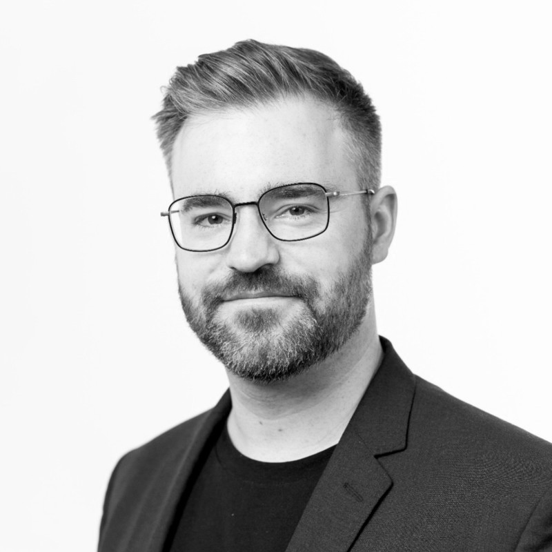 A black and white headshot of a man with short hair, glasses, a beard, and a mustache, wearing a dark blazer and a T-shirt, smiling slightly and looking at the camera.