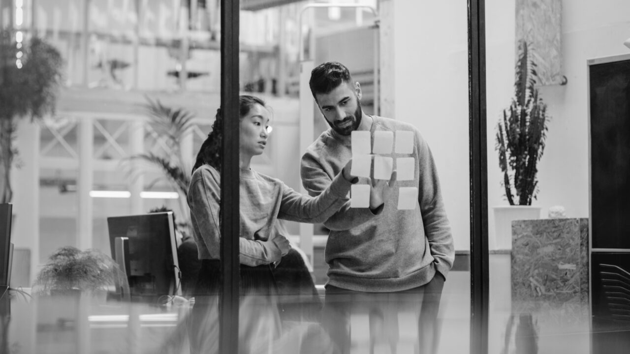Two people in an office looking at sticky notes on a glass wall, seemingly discussing or planning something. Both are dressed in casual attire.