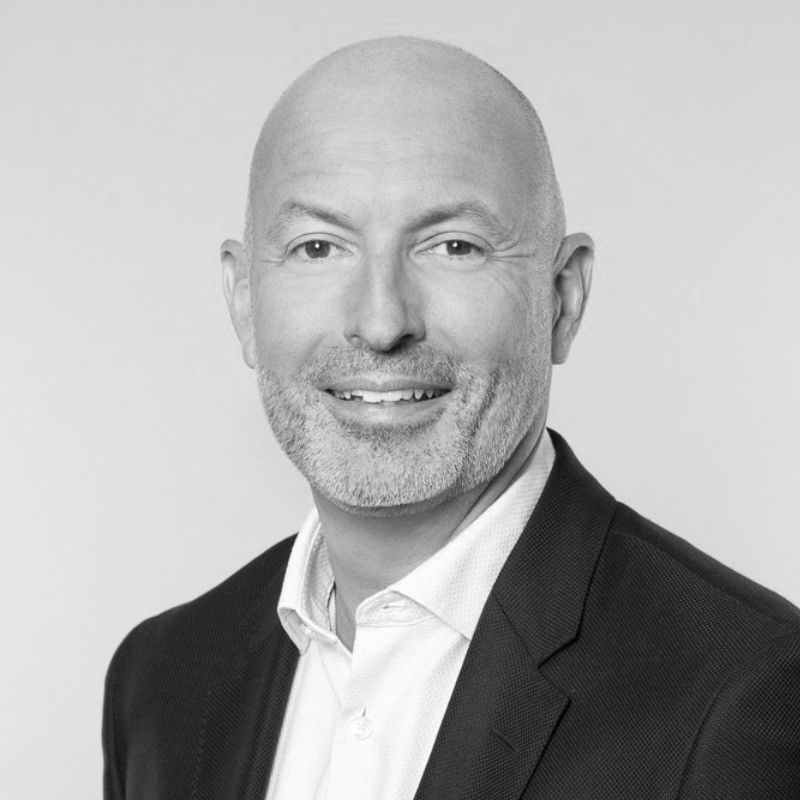 A bald man with a short beard, dressed in a dark blazer and white shirt, is smiling in this black and white portrait.