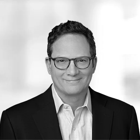 A man wearing glasses and a suit jacket over a collared shirt smiles slightly. He stands against a blurred, light-colored background.