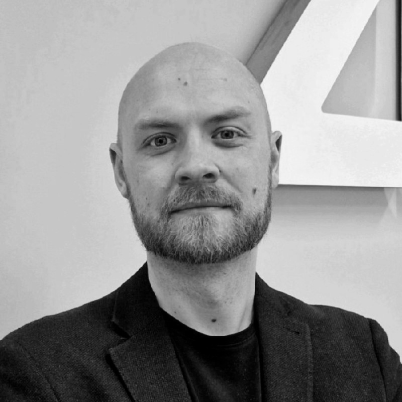 A black-and-white portrait of a bald man with a beard, dressed in a blazer and t-shirt, standing in front of a white wall featuring part of a large, geometric number 4.