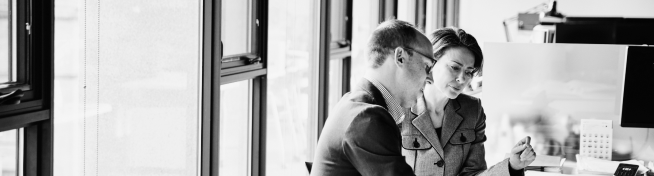 Two people in business attire discuss something while standing near office windows, with desks and equipment in the background.