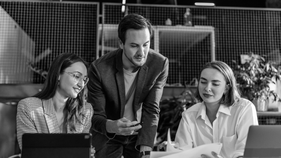 Three colleagues in a discussion at a workspace. A man stands between two seated women, conversing and reviewing documents.