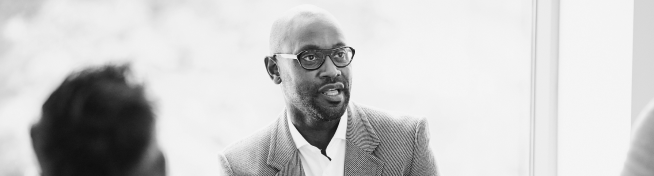 A man wearing glasses and a suit jacket speaks during a discussion, with blurred figures in the foreground.