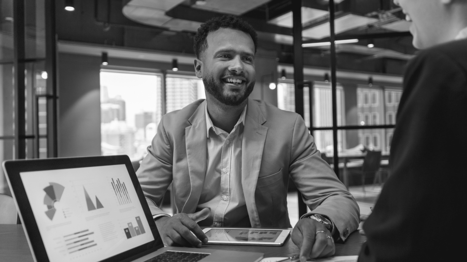 A man in a suit is smiling during a conversation in a modern office. He is sitting at a table with graphs displayed on a laptop and holding a tablet.