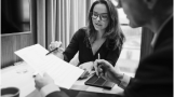 A woman and a man in formal attire review a document together at a desk, with a tablet and cup nearby.