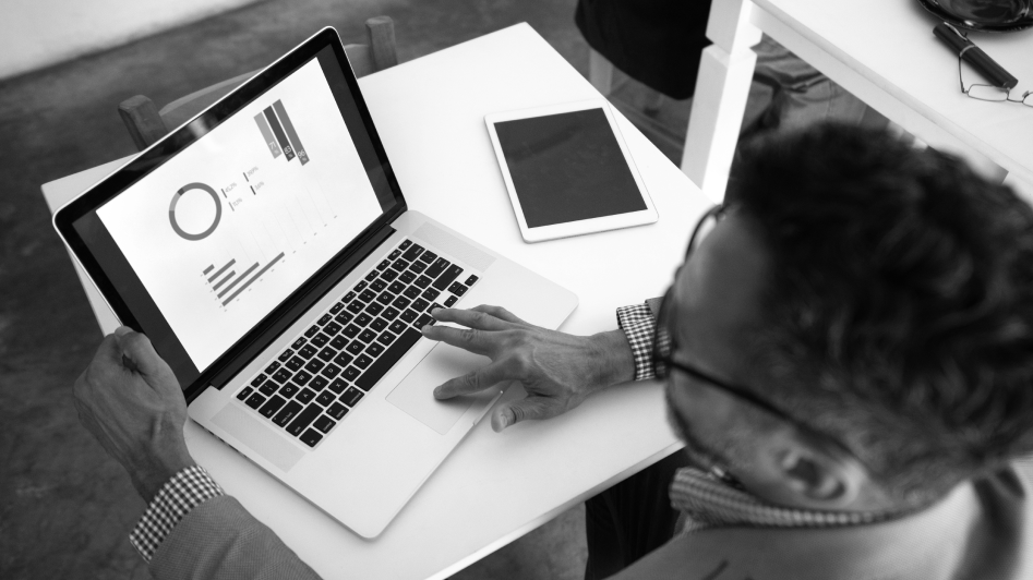 A person is viewing data visualizations on a laptop screen at a white desk. A tablet and a pair of glasses are also on the desk.