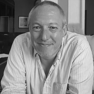 In a black and white photo, a man with short hair and a beard smiles while wearing a striped shirt at a Zuora Subscribed Live event indoors.