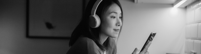 A woman wearing headphones is looking at her smartphone in a dimly lit room.