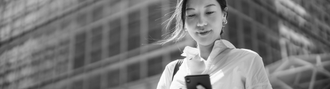 A woman stands outdoors, glancing at her smartphone, with a city building in the background. The image is in black and white.