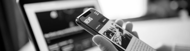 A person holds a smartphone displaying a news article, with a laptop screen in the background. Image is in black and white.