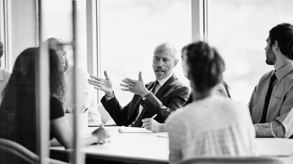 A man in a suit gestures while speaking during a meeting with four other people in an office conference room.