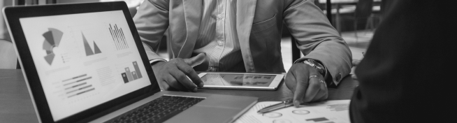 Two professionals discuss data charts and graphs on a laptop and tablet during a meeting.