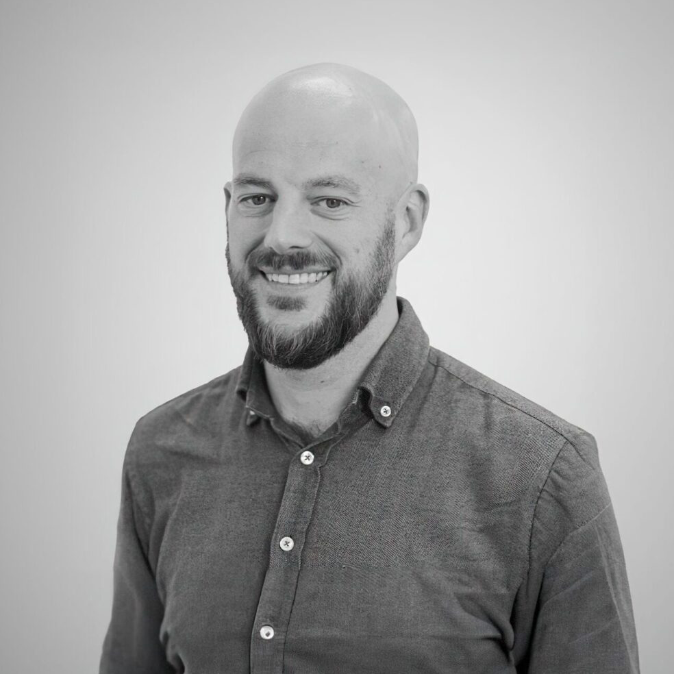 A bald man with a beard, wearing a button-up shirt, smiles at the camera against a plain background.