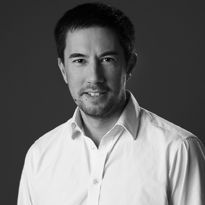 A black-and-white portrait of a man with short dark hair, wearing a buttoned-up white shirt, smiling slightly, against a plain dark background, embodies the focus and energy often seen at Zuora Subscribed Live events.