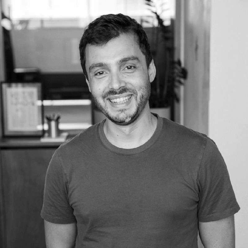 A man with short hair and a beard smiles while standing in a room with blurred background elements. He is wearing a casual, short-sleeved T-shirt.