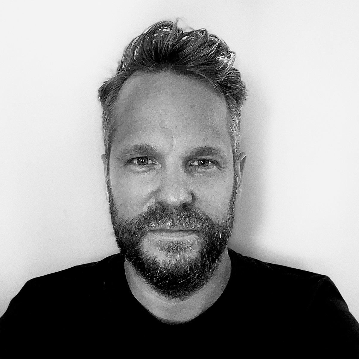 A black-and-white portrait of a man with short hair and a beard, wearing a dark shirt, looking directly at the camera against a plain background.