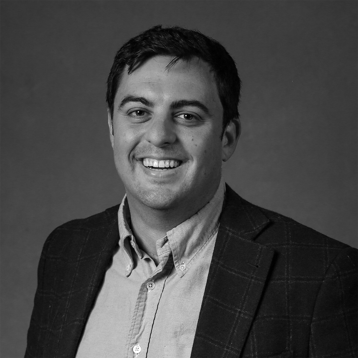 Black and white portrait of a man smiling, wearing a light-colored shirt and a dark blazer, against a plain background.