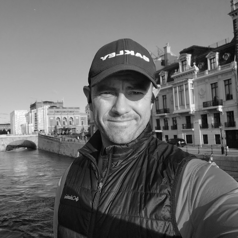 A person wearing a cap and jacket stands on a bridge. Buildings and a river are visible in the background. The image is in black and white.