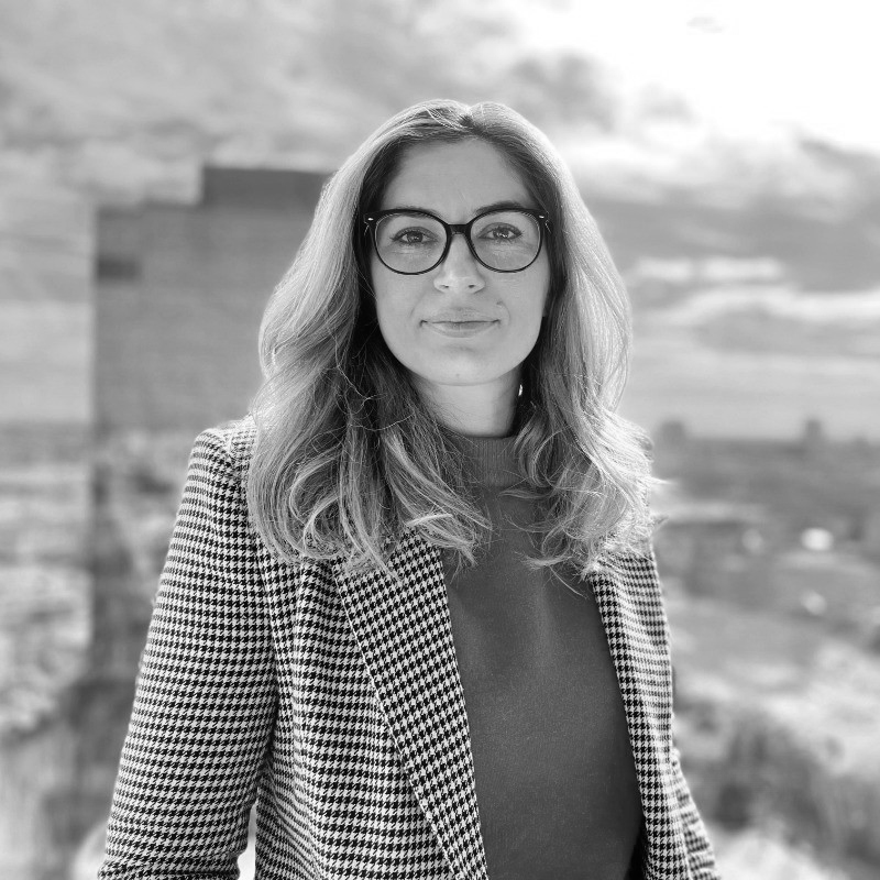 A woman with long hair and glasses stands confidently outdoors, wearing a houndstooth-patterned jacket and a plain top. The background features an urban scene with buildings and a cloudy sky.