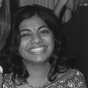 A smiling person with shoulder-length hair and wearing a traditional outfit with a bindi, shown in a close-up black and white photograph.