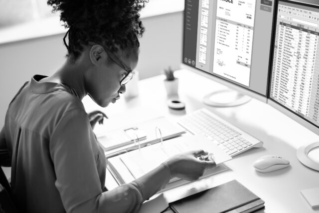 A person wearing glasses works at a desk, examining documents while two computer screens display an invoice and a spreadsheet.